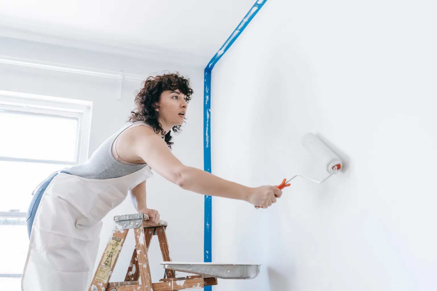 A house painter mid-job at a residential site, working with a roller pole and cut-in brush, hands and work in sharp focus, the surrounding room softly out of focus